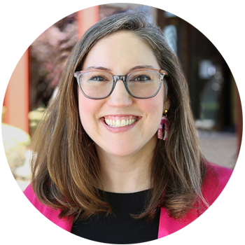 Portrait of a smiling woman with long brown hair and glasses, wearing a pink blazer.
