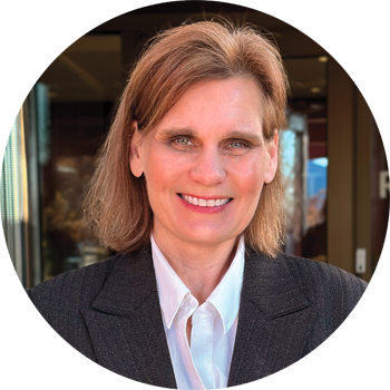 A professional headshot of a woman with medium-length brown hair, wearing a gray blazer and a white shirt, smiling in front of a neutral background.