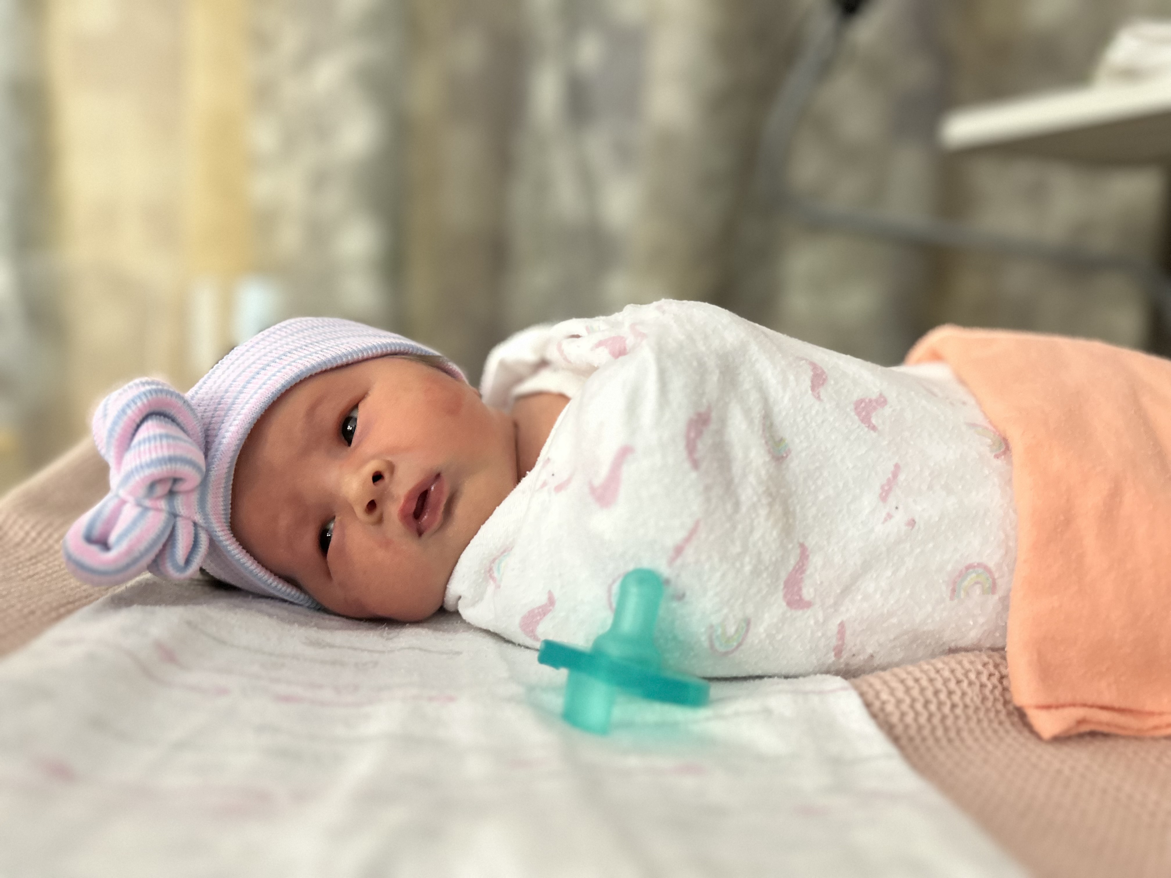 A newborn baby lying on a blanket, wearing a light purple hat with a bow and swaddled in a white blanket with pink patterns, beside a pacifier.