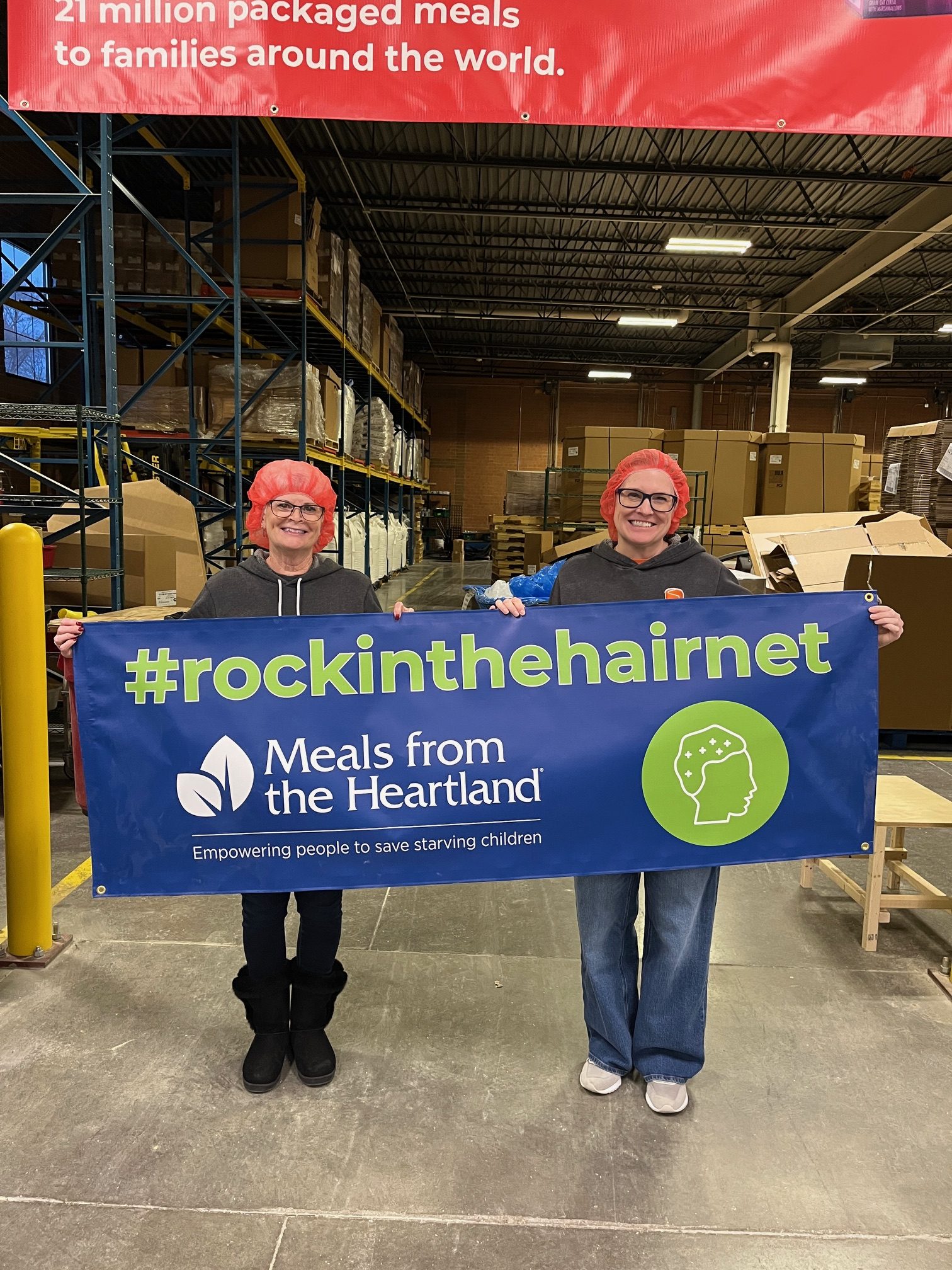 Two women wearing red hairnets hold a large banner promoting the '#rockinthehairnet' campaign for Meals from the Heartland, inside a food packaging facility.